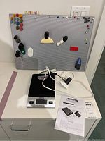 Photo of tack board with assorted thumb tacks, clips, magnets on a white surface beside black and silver digital postal scale and cables with paper manual.