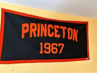 Large rectangular Princeton 1967 felt banner hanging on a wall, showing orange letters on black background with red border.