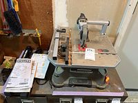 Rockwell BladeRunner RK7320 saw on a workbench showing attachment arms and the saw blade. The saw is dusty, indicating it needs cleaning and has not been recently used.