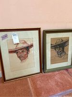 Two framed original art portraits leaning on a wall, showing two men wearing hats.