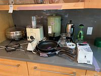 Full view of small kitchen appliances and cookware including FarberWare electric fry pan, frying pans, food processor, hand mixer, coffee pots, green container, and kitchen tools