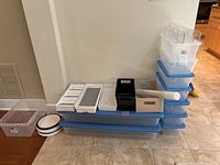 Stack of plastic storage bins in clear and blue with lids, smaller organizing trays and lazy Susans on tiled floor against wall.