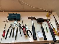Wide shot of the tool collection including Makita drill set, hammers, oil can, pliers, screwdrivers, and file on a beige table.