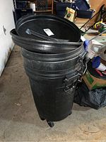 Photo showing one Rubbermaid black trash can with lid resting on top. The surface appears scratched and dusty.