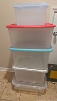 Stack of five translucent plastic storage bins of varying sizes and lid colors on tiled floor.