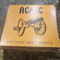 Top view of AC/DC 'For Those About to Rock' vinyl record cover, tan with image of cannon and bold AC/DC logo.