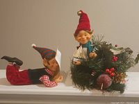 Pair of Christmas elves, one lying holding scroll and the other with a wreath and miniature gifts.