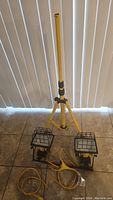 View showing the yellow tripod stand extended upright on tiled floor with two construction lights placed at the bottom, cords visible.