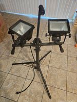 Two black metal construction lights mounted on a black metal tripod stand positioned on a floor. Both lights have glass fronts and reflective interiors, power cords visible.