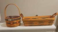 Two vintage Longaberger baskets placed side by side on a white surface against a plain background. One is round with a handle and a fabric liner, the other is rectangular with two side handles and no liner.