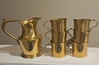 Brass pitcher placed next to six stacked brass cups showing the entire set