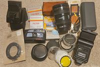 Assortment of vintage camera accessories including lenses, lens caps, flash units, remote control, filter boxes, and film canisters.