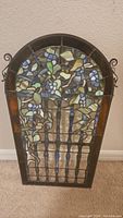 Full view of the vintage leaded stained glass window panel depicting a blue floral design with green leaves, arched top shape, and basket/fence vertical elements at the bottom. Shows overall condition and colors.