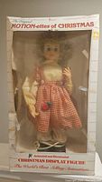 Front view of the boxed Christmas animated display figure showing the figure in a checkered dress with curly blonde hair and a rose detail.