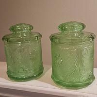 Two green glass canisters side by side showing the raised Chantilly floral and scroll design details.