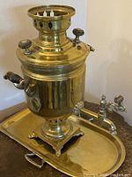 Front view of the brass samovar placed on the brass tray, showing the main body, top chimney with ventilation holes, and side wooden handle on the right.
