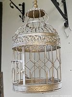 Front and side angle view of white metal decorative bird cage with hinged door, scrollwork, and hanging hook