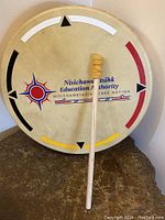 Front view of Indigenous drum with attached wooden drum stick leaning on it, showing the logo and colored arrow designs on the drum head.
