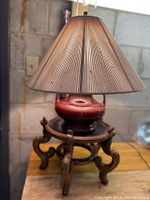 Table lamp with ribbed brownish lampshade over red ceramic lamp body resting on wooden stand.