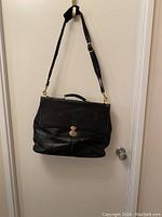 Front view of black leather messenger bag hanging, showing flap with gold-tone twist lock and adjustable shoulder strap.