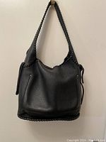 Front view of the black leather handbag hanging against a wall, showing braided shoulder strap and soft handbag structure.