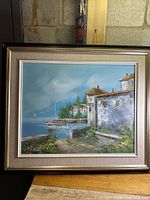Framed original oil painting showing a scenic coastal village with boats docked at the pier and white buildings with tiled roofs, under a blue sky.