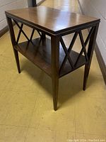 Angled front view of vintage Sheraton style mahogany side table showing rectangular top, lower shelf, and crossed inlay patterns on sides, on a tiled floor with a hallway background.