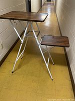 Side angled photo of vintage folding projector table showing larger top and smaller side shelf with white metal folding legs on yellow floor in hallway.