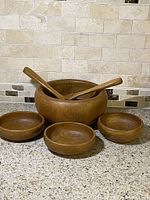 Front view of the large wooden bowl surrounded by three smaller bowls and two utensils on granite countertop with tiled backsplash.