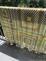 Close-up photo of the vintage hand crochet bedspread draped over railing, showing large granny square crochet pattern in shades of green, yellow, and white.