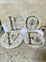 Four porcelain plates arranged to spell the word LOVE with ornate baroque styling and cherub illustrations.