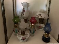 Photo shows six antique miniature oil lamps with various styled glass chimneys and bases decorated with floral designs. Brass burner parts visible. The lamps are placed on a shelf against a wall.
