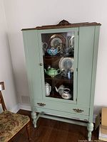Front view of antique sage green painted hutch with glass door showing interior shelves with china and decorative plates.