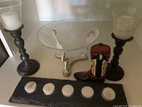 Two tall candleholders with large white pillar candles, slate tray with five white round tealight candles, clear decorative glass bowl on white stand, small red and black cowboy boot figurine.