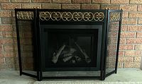 Front view of the black foldable metal fireplace screen with decorative scrollwork trim, placed in front of a brick fireplace, showing the overall condition and design.