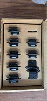 Box containing 8 sets of black metal hinges with mounting brackets arranged inside