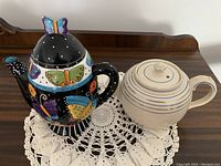 Two teapots placed side by side on a wooden surface with a white crochet doily underneath. One teapot is colorful with butterfly designs, the other is cream with blue rings.