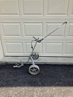 Silver three-wheeled pull golf cart with a long handle, placed in front of a closed garage door, showing side profile and front view.