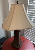 Full lamp showing beige fabric shade and wooden base with nailhead trim on a red table beside a window with blinds.
