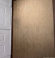 Top-down photo showing light brown jute rug with chocolate trim placed in a hallway or room with white doors on the side.