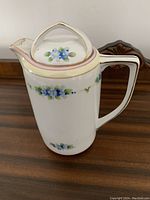 Full side view of tall white porcelain teapot with blue floral decoration, pink and gold trim on spout, handle, lid and body.