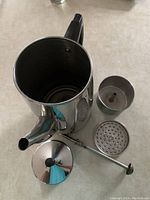 Top view of disassembled stainless steel 10-cup percolator showing lid, coffee basket, perforated plate, stem, and body with black handle and spout.