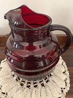 Side angle view showing deep red color, ribbed design, handle and wide spout on large glass jug.