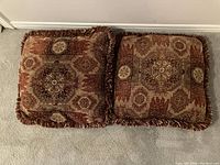 Two square throw pillows with ornate brown and beige geometric pattern and brown fringed edges, laid out to show patterned side.