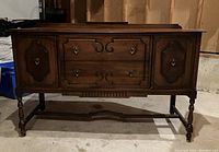 Front view of vintage solid wood buffet showing two cupboard doors with decorative panels, two drawers with ring pulls, and spindle legs with stretcher.
