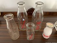 Five vintage glass milk bottles of varying sizes placed on wooden surface. One sealed with cap, one with chipped spout, others with red logos.
