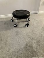 Black low work stool on wheels with round padded seat and chrome legs, positioned on carpeted floor.