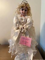 Porcelain doll named Angelina, dressed as a white princess with crown and long white dress, holding flowers, standing upright.