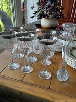 6 crystal stem glasses with wide silver rims and one tall clear crystal bud vase on wooden table near window.