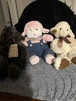 Three small stuffed lamb toys displayed on a gray surface, photographed against a black backdrop, showing different plush designs and colors.